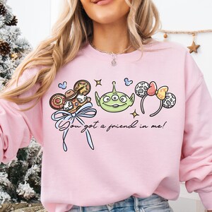 A woman wearing a pink sweatshirt with a graphic design featuring a green alien character and various Disney-themed accessories.