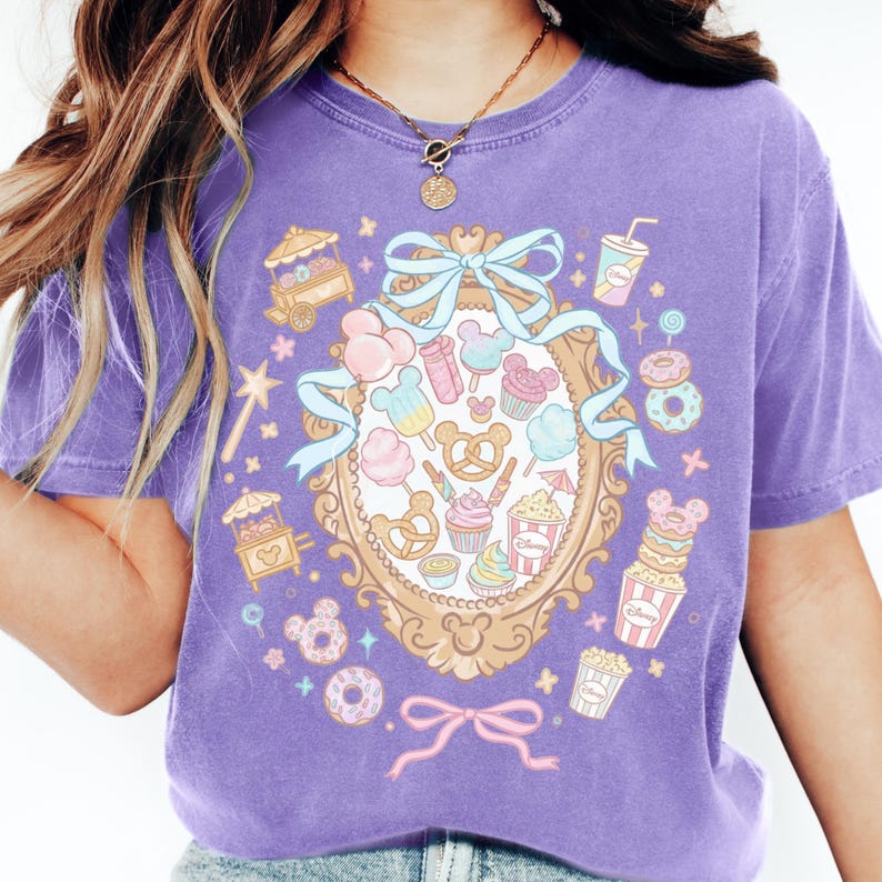 A person wearing a purple t-shirt with a colorful graphic design featuring various Disney-themed items such as Mickey Mouse, popcorn, and candy.