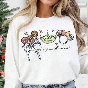 A woman wearing a white sweatshirt with a graphic design featuring a green alien character and various Disney-themed elements.