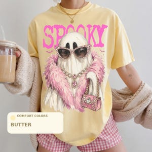 A person wearing a yellow t-shirt with a graphic of a ghost wearing sunglasses and a pink feather boa, holding a drink.
