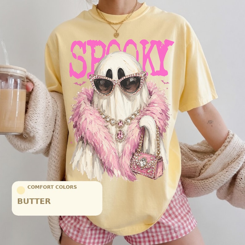 A person wearing a yellow t-shirt with a graphic of a ghost wearing sunglasses and a pink feather boa, holding a drink.