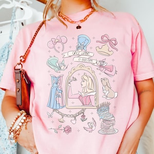 May include: Pink t-shirt featuring a graphic of Sleeping Beauty and related imagery. The design includes a framed portrait, a dress, a crown, and the text "ONCE UPON A DREAM". Other elements include a bunny, a bird, and a cake.