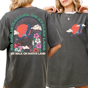 May include: Dark gray t-shirts with a graphic design featuring a red sun, a flying bird, and flowers. The text on the back of the shirt reads "NO ONE IS ILLEGAL ON STOLEN LAND" and "WE WALK ON NATIVE LAND".