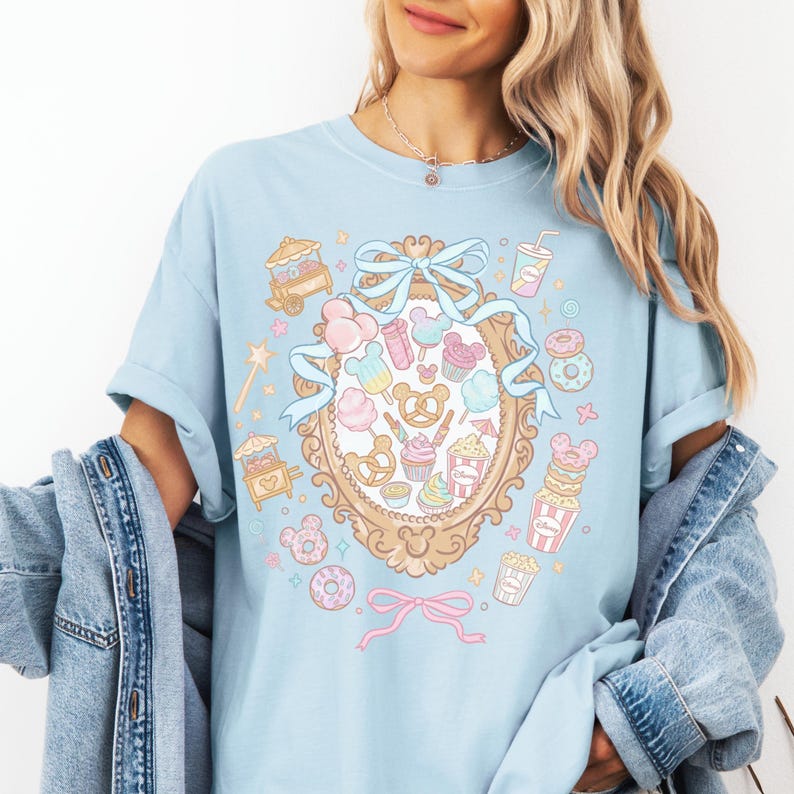 A woman wearing a light blue t-shirt with a whimsical graphic design featuring various cartoon-like objects and characters, such as donuts, Mickey Mouse, and a mirror.