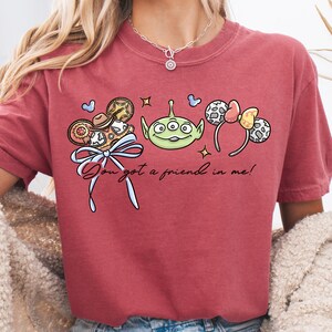 A person wearing a red t-shirt with a graphic design featuring a green alien character, a bow, and other cartoon-style elements.