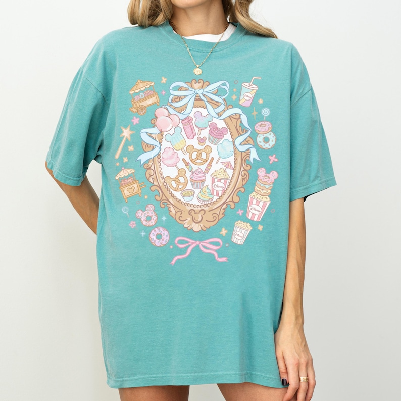 A woman wearing a turquoise t-shirt with a colorful graphic design featuring various cartoon-like elements such as donuts, cupcakes, and a mirror.