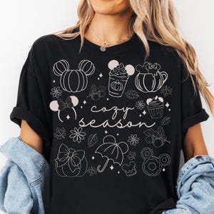 A woman wearing a black t-shirt with a graphic design featuring various Disney-themed illustrations, including Mickey Mouse, pumpkins, and umbrellas, along with the text "cozy season".