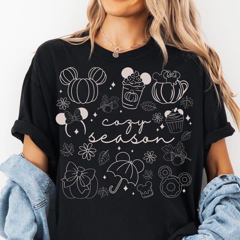 A woman wearing a black t-shirt with a graphic design featuring various Disney-themed illustrations, including Mickey Mouse, pumpkins, and umbrellas, along with the text "cozy season".