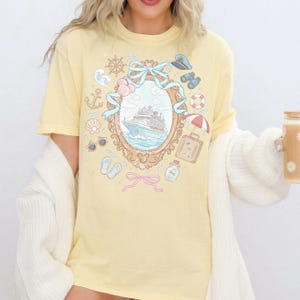 A woman wearing a yellow t-shirt with a ship design and holding a drink.