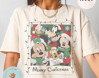May include: Ivory-colored t-shirt featuring a Christmas-themed graphic of Disney characters, including Mickey and Minnie Mouse, Donald Duck, and Goofy. The graphic is framed with a string of lights and the words "Merry Christmas".