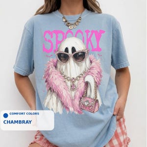 A person wearing a blue t-shirt with a graphic design of a ghost wearing sunglasses, a pink feathered scarf, and holding a pink purse.