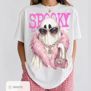 A white t-shirt with a graphic design of a ghost wearing sunglasses, a pink feathered scarf, and holding a pink purse.