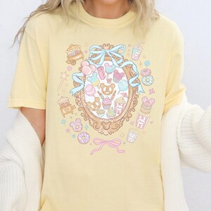 A person wearing a yellow t-shirt with a colorful graphic design featuring various cartoon-like objects and characters, such as donuts, cupcakes, and Mickey Mouse.