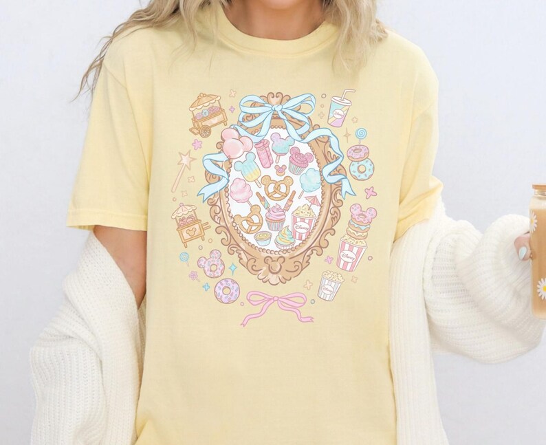 A person wearing a yellow t-shirt with a colorful graphic design featuring various cartoon-like objects and characters, such as donuts, cupcakes, and Mickey Mouse.