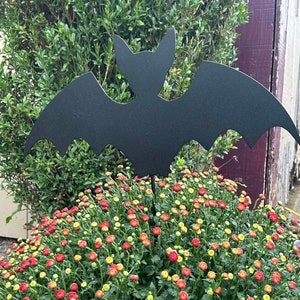 May include: A black metal bat silhouette garden stake. The bat is positioned above a vibrant bush of red and yellow mums. The background features green foliage and a glimpse of a wooden structure, creating a seasonal, Halloween-themed display.