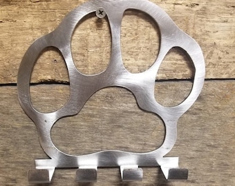 May include: A silver metal paw print key holder with five hooks. The paw print is attached to a wooden surface with a screw.