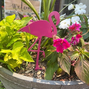 May include: A bright pink metal flamingo garden stake stands in a large gray planter filled with green and red plants and pink and white flowers. A welcome sign is visible in the background.