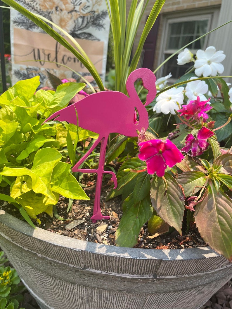 May include: A bright pink metal flamingo garden stake stands in a large gray planter filled with green and red plants and pink and white flowers. A welcome sign is visible in the background.