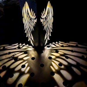 May include: A dark, rectangular sculpture with two illuminated wing designs. The wings are detailed with a layered feather pattern, casting intricate shadows on the surface below. The sculpture is set against a dark background.