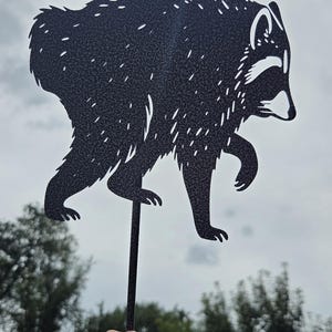 May include: A black metal silhouette of a raccoon with cut-out details representing fur texture and facial markings. The raccoon figure is attached to a thin, straight metal stake extending downward, designed for placement in a garden or outdoor space.