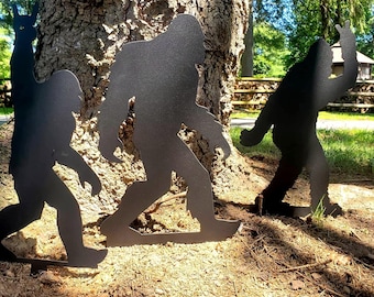 May include: Three black metal Bigfoot yard art silhouettes are displayed in front of a tree. One silhouette is making a rock on hand gesture, another is making a peace sign, and the third is walking.