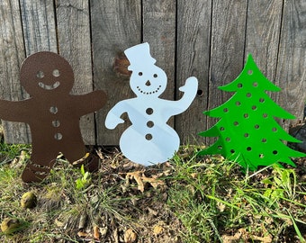 May include: Three metal Christmas yard decorations: a gingerbread person, a snowman, and a Christmas tree. The gingerbread person is brown, the snowman is white, and the Christmas tree is green.