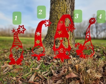 May include: Four red metal gnomes with white snowflake accents and a white star on their hats. The gnomes are standing in a grassy area with brown leaves.