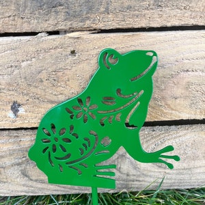 May include: A green metal frog with a floral design, standing on a green metal stake. The frog is facing right and has a happy expression.