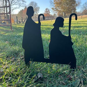 May include: Two black metal silhouettes of shepherds with staffs standing in a grassy field. The shepherds are facing each other and are in a kneeling position.