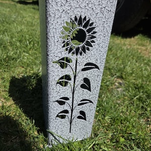 May include: A tall, rectangular metal garden stake featuring a textured, crackled gray and white finish. The front surface displays a laser-cut silhouette of a sunflower with a central bloom and a stem adorned with several pointed leaves.
