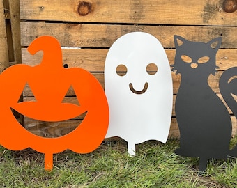 Puede incluir: Tres decoraciones de jardín de Halloween: una calabaza naranja con una cara de calabaza tallada, un fantasma blanco con una cara sonriente y un gato negro con una cola enroscada.