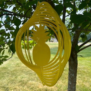 May include: A yellow metal pineapple wind chime with a circular design. The chime is hanging from a tree branch.