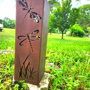 May include: A rectangular, bronze-colored metal garden stake with dragonfly cutouts. The stake features two dragonfly silhouettes and grass-like cutouts near the base. It is set on a stone in a grassy area.