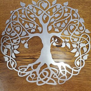 Op de afbeelding: Een zilveren metalen wanddecoratie in de vorm van een levensboom met ingewikkelde takken en wortels. De boom is omringd door bladeren en heeft een hartvorm aan de bovenkant.