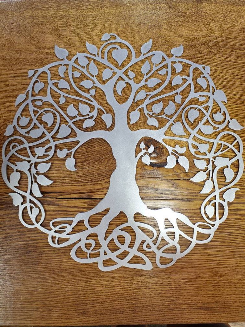 Op de afbeelding: Een zilveren metalen wanddecoratie in de vorm van een levensboom met ingewikkelde takken en wortels. De boom is omringd door bladeren en heeft een hartvorm aan de bovenkant.