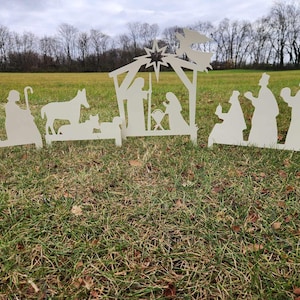 May include: White silhouette cutout of a nativity scene with a star, angel, and figures of Mary, Joseph, and baby Jesus. The scene is set up on a grassy lawn.