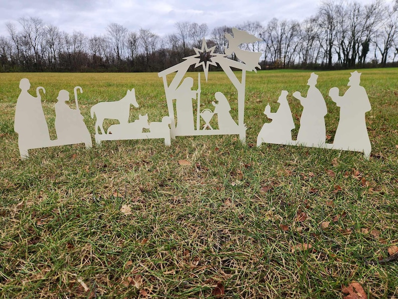 May include: White silhouette cutout of a nativity scene with a star, angel, and figures of Mary, Joseph, and baby Jesus. The scene is set up on a grassy lawn.