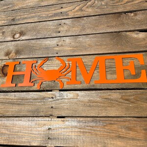 May include: Orange metal HOME sign with a crab silhouette in the center. The sign is on a wooden surface.