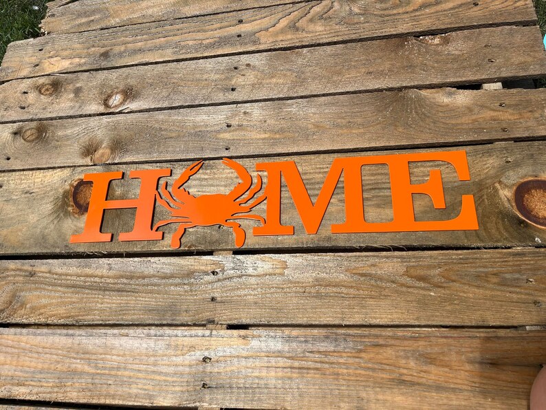 May include: Orange metal HOME sign with a crab silhouette in the center. The sign is on a wooden surface.