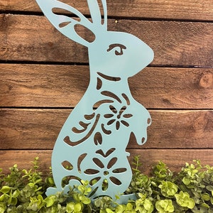 May include: A light blue metal bunny cutout with a floral design. The bunny is standing in front of a wooden background and green foliage.