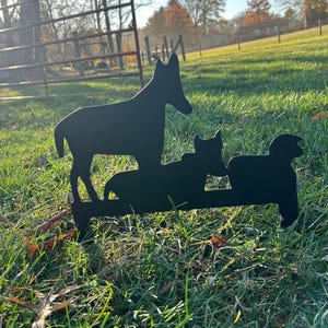 May include: Black metal silhouette of a dog and two cats standing on a grassy lawn. The dog is standing upright with its head tilted to the side, while the cats are sitting down. The silhouette is a yard decoration.