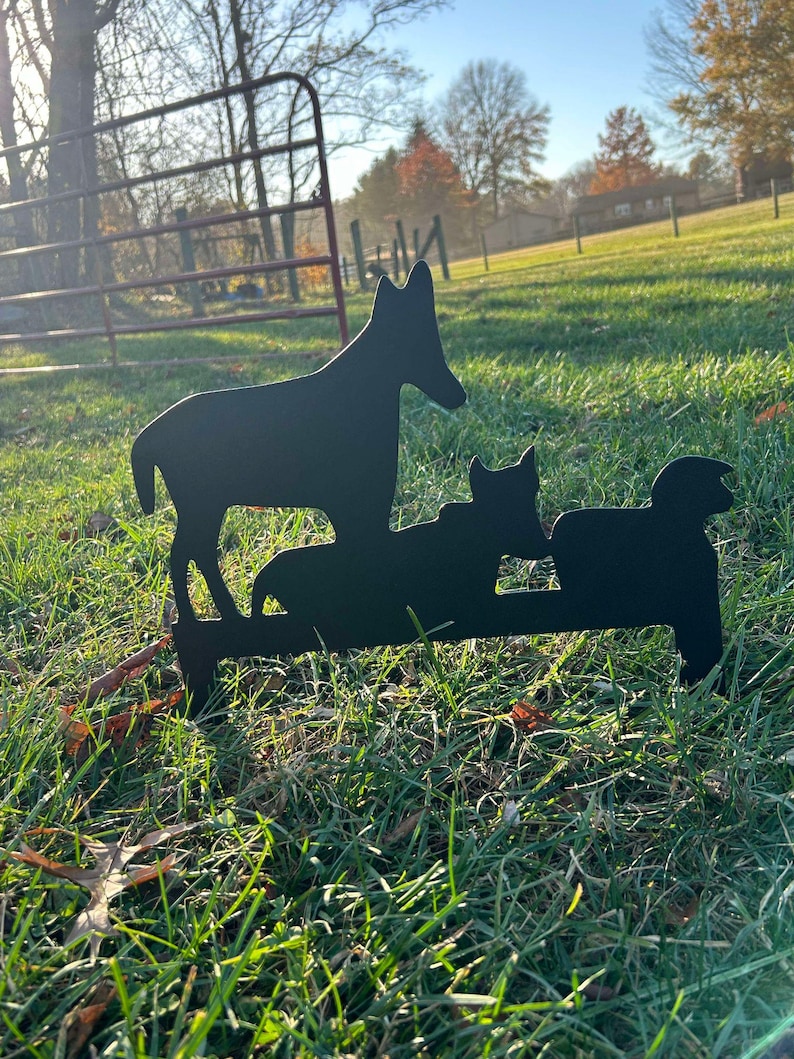 May include: Black metal silhouette of a dog and two cats standing on a grassy lawn. The dog is standing upright with its head tilted to the side, while the cats are sitting down. The silhouette is a yard decoration.
