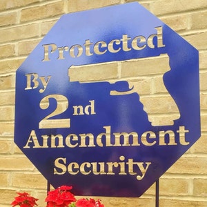 May include: A blue metal sign with the text "Protected By 2nd Amendment Security" in gold lettering. A silhouette of a handgun is in the center of the sign.