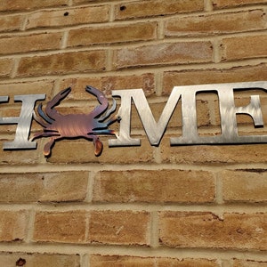 May include: Metal wall art featuring the word "HOME" with a stylized crab in the center. The letters are silver and the crab is a reddish-brown color.