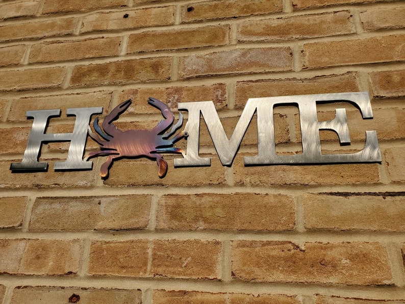May include: Metal wall art featuring the word "HOME" with a stylized crab in the center. The letters are silver and the crab is a reddish-brown color.
