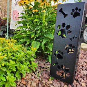 May include: A black metal pet paw print garden stake. The rectangular stake features cut-out paw prints and bone shapes. The stake is placed in a garden bed with red rocks and green plants, with yellow flowers in the background.