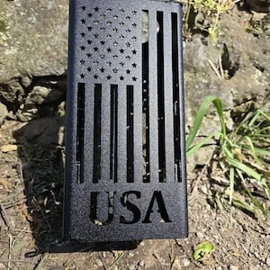 May include: A rectangular black metal cover featuring a laser-cut design of the American flag with stars and stripes. The letters USA are cut out at the bottom of the panel. The object has a matte finish and a vertical orientation with open slots.