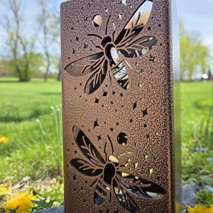 May include: A rectangular metal lantern with a textured bronze finish featuring laser-cut designs of two bees surrounded by small stars and circular shapes. The structure has open cutouts that allow light to pass through the bee and star patterns.