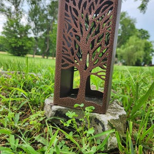 May include: A rectangular metal lantern with a textured bronze finish featuring a laser-cut tree silhouette on the front panel. The structure is open at the top and bottom, showcasing a dark interior surface and a sturdy, hollow frame design.