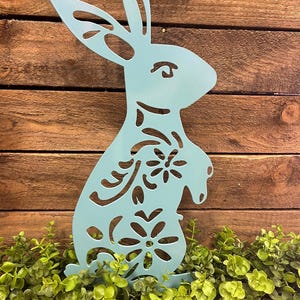 May include: A light blue metal Easter bunny silhouette with floral cutouts. The bunny is standing upright with its ears perked up. The bunny is placed in front of a wooden background and green foliage.